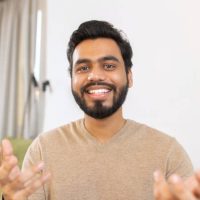 Portrait of happy young Indian guy sitting at desk and gesturing. Smiling ethnic man involved virtual meeting. Hispanic male student taking a part in educational conference